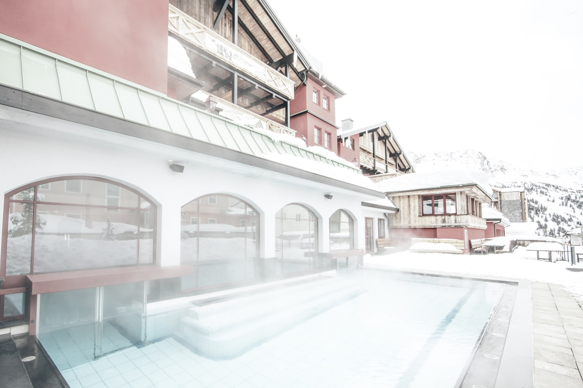 RR_Bild-03_RGB_Wellness Eine ruhige, verschneite Szene in einem Gebirgsort, wo ein dampfendes Freibad einen warmen Kontrast zur kühlen Umgebung bietet.