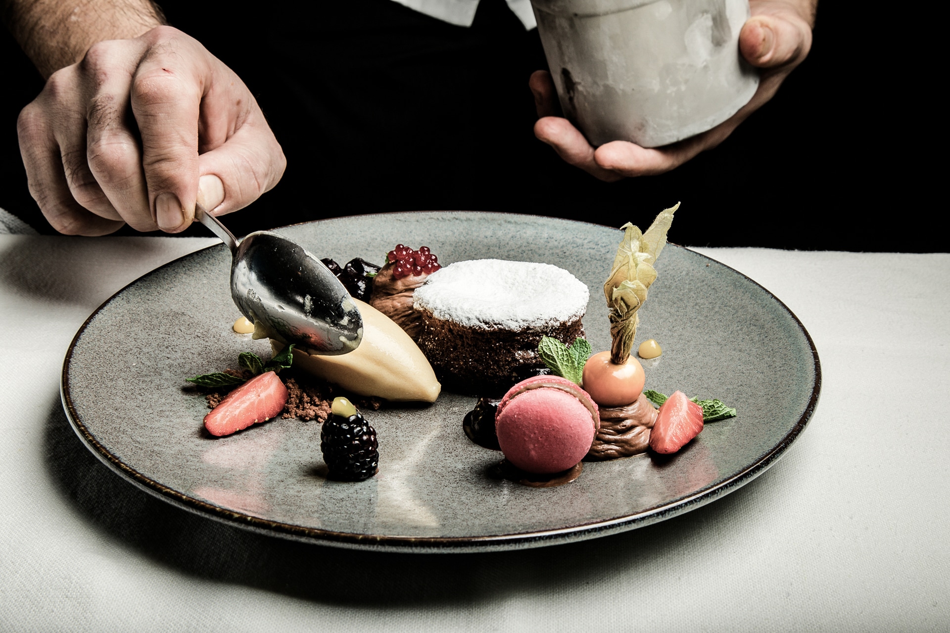 rigele-royal-kulinarik-01 Ein Koch, der eine Gourmet-Dessertplatte mit delikater Schokoladensoße veredelt und ein kunstvolles Arrangement aus einem Schokoladenkuchen, einer Eiscreme, Macarons und frischen Beeren präsentiert.