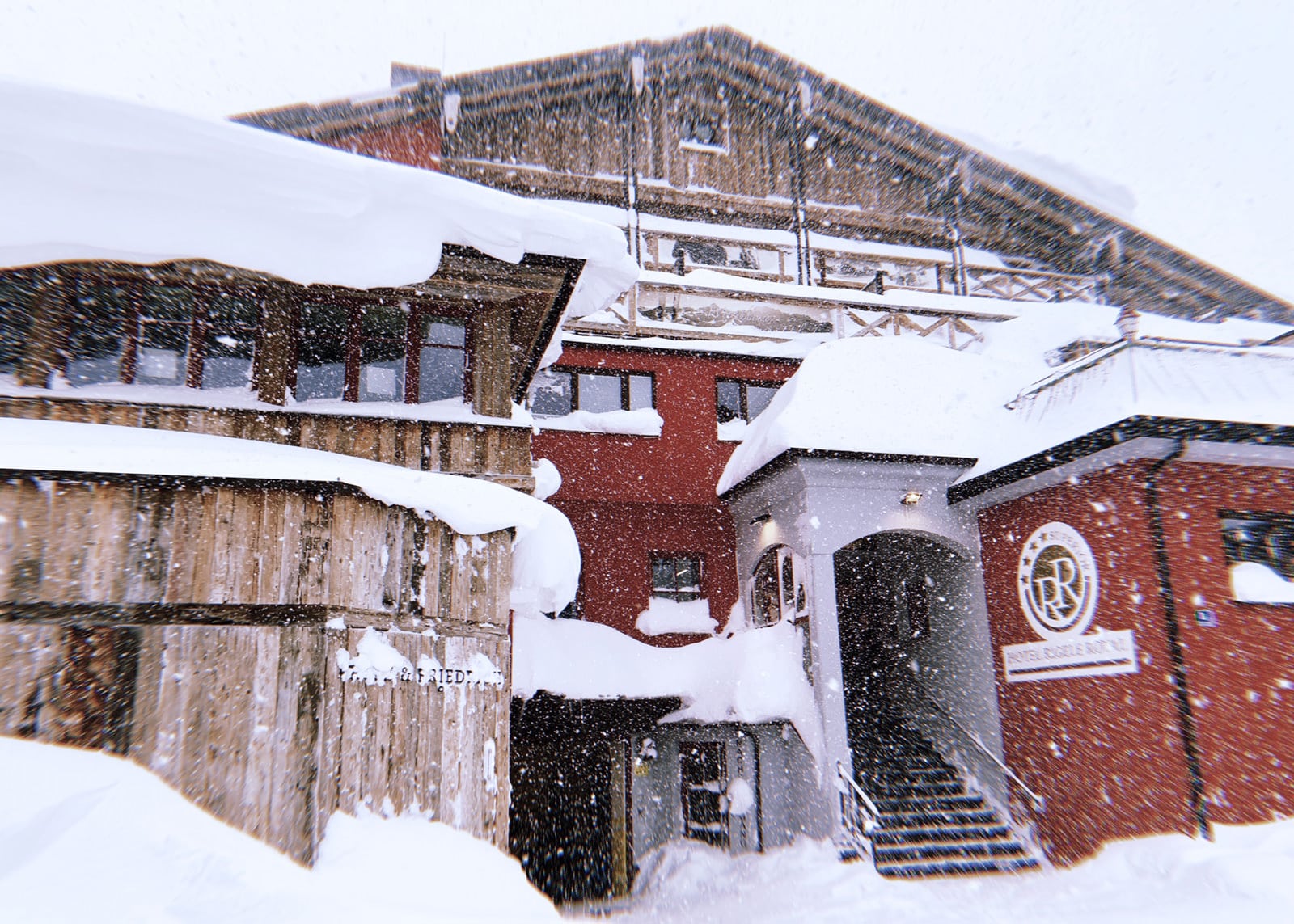 hotel-aussen-schnee Eine bezaubernde Winterszene, die schneebedeckte Gebäude in einem verschneiten Dorf zeigt, mit großen Verwehungen, die sich auf den Dächern absetzen, und einem heiteren Schneegestöber, das vom Himmel fällt.