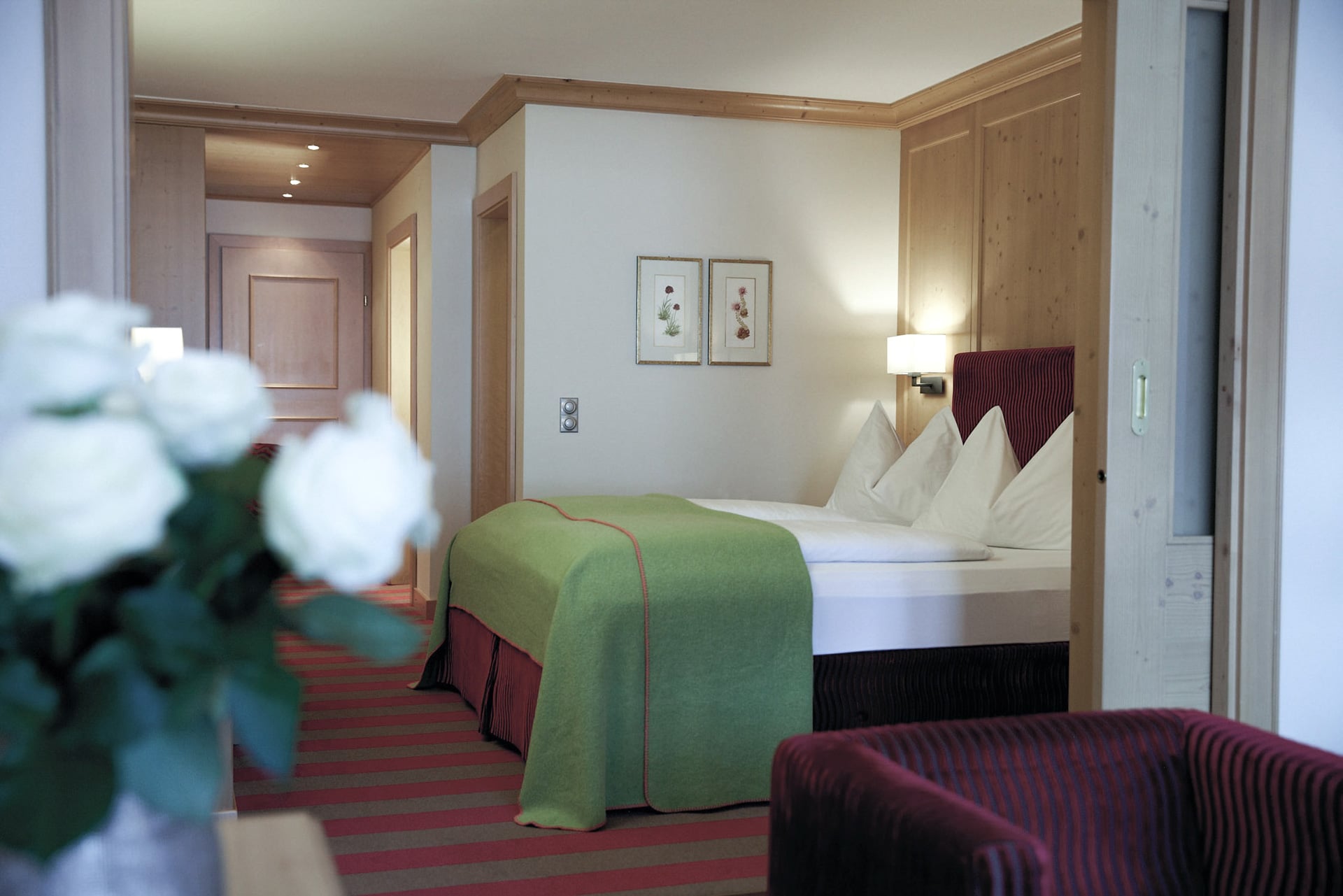 junior-suite-deluxe-sauna-01 Ein gemütliches Hotelzimmer mit einem ordentlich gemachten Bett, das mit weißen Kissen und einer grünen Decke geschmückt ist. Das Zimmer verfügt über helle Holzwände, einen kleinen Flur, der zum Eingang führt, zwei gerahmte Bilder über dem Bett und eine Vase mit weißen Blumen im Vordergrund.