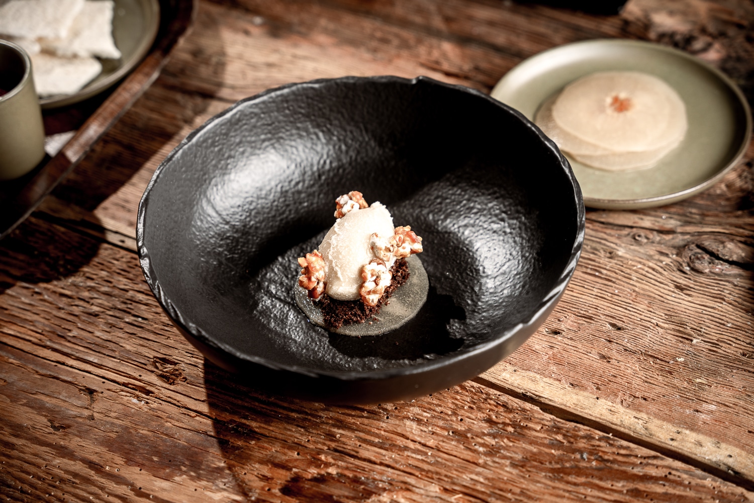 DSC05933 Ein Gorumet-Dessert mit dunklem Teig, einer Mousse darauf und mit Walnüssen garniert, serviert in einer schwarzen Schüssel auf einer rustikalen Holz-Tischplatte